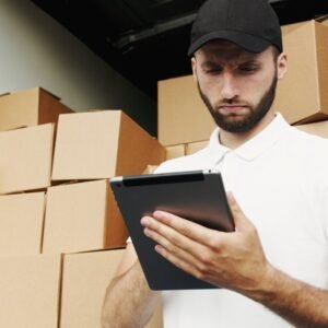 Delivery worker using a tablet to manage shipments with stacked boxes in the background.